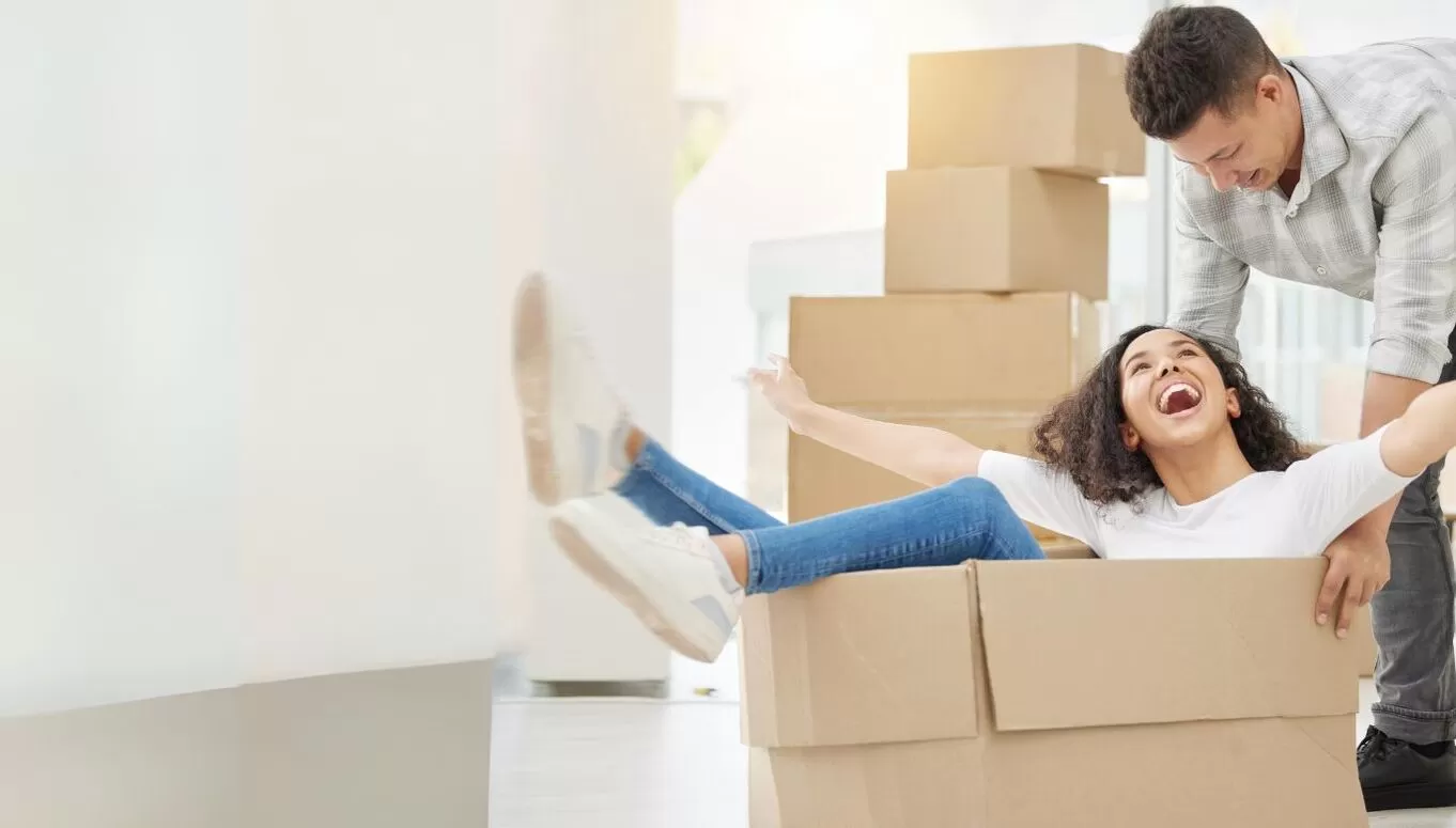 A man pushing a woman sitting in a cardboard box, both smiling and having fun during a moving day.