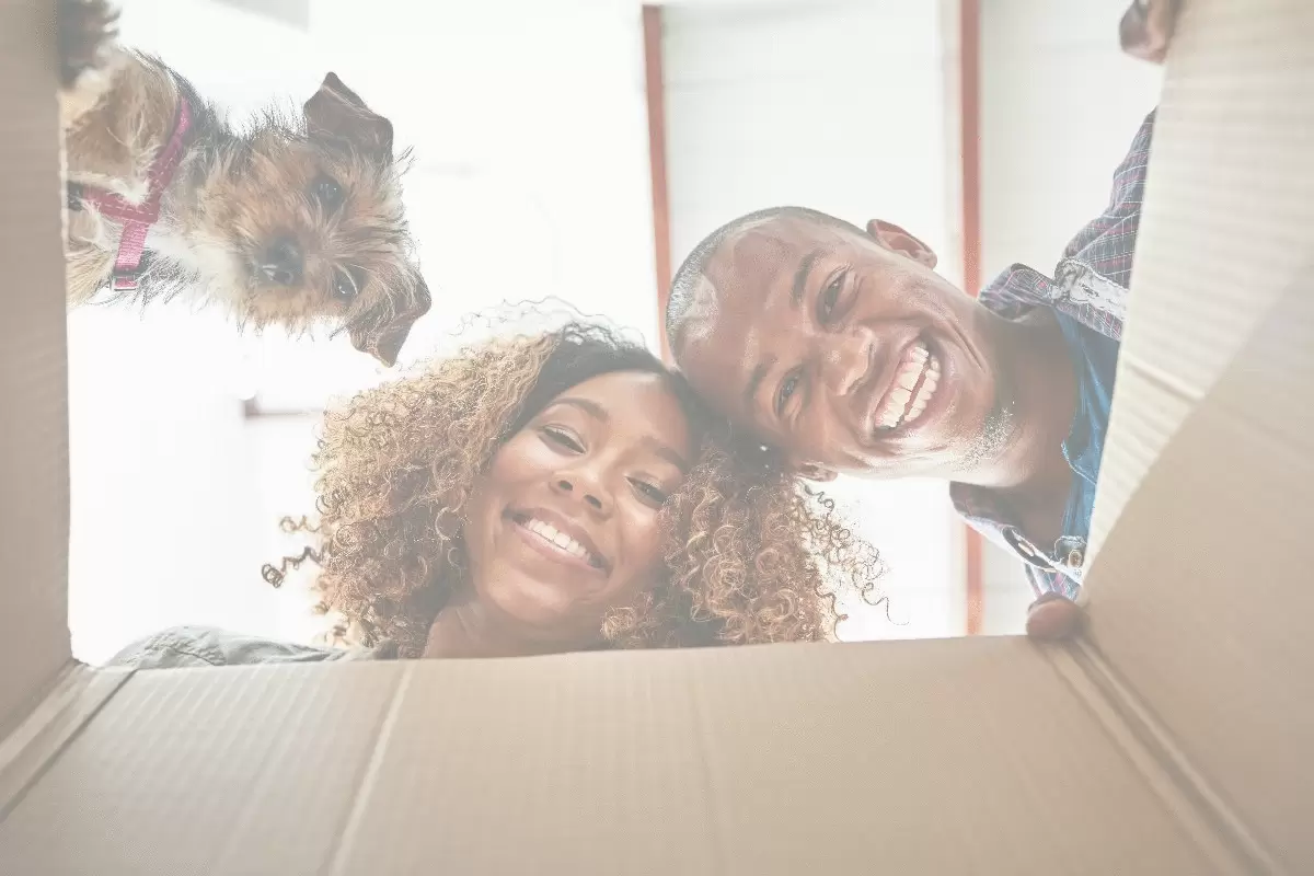 A smiling couple peeking into an open box along with their playful dog.